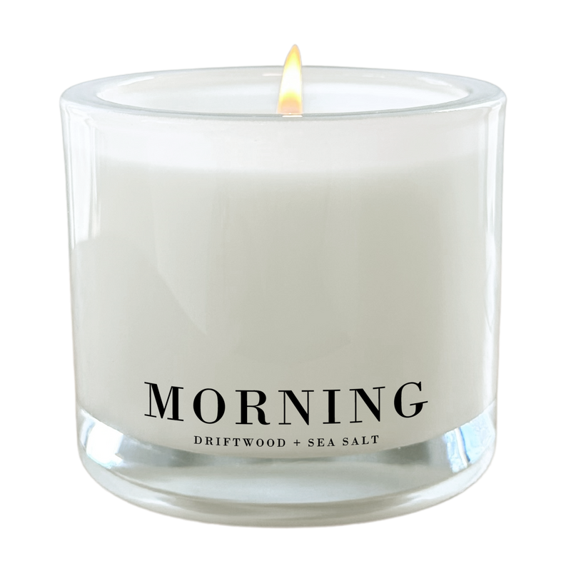 A white candle with a lit flame, labeled as "MORNING" and "Driftwood + Sea Salt".