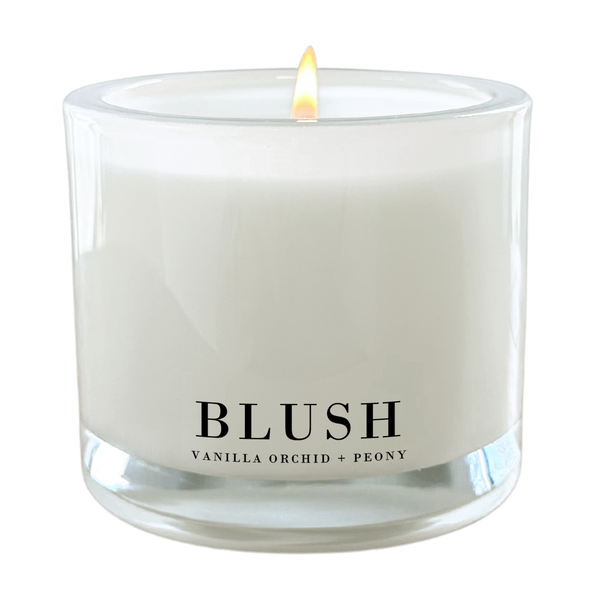 A white candle with a lit flame, labeled as "BLUSH" and containing vanilla orchid and peony scents.