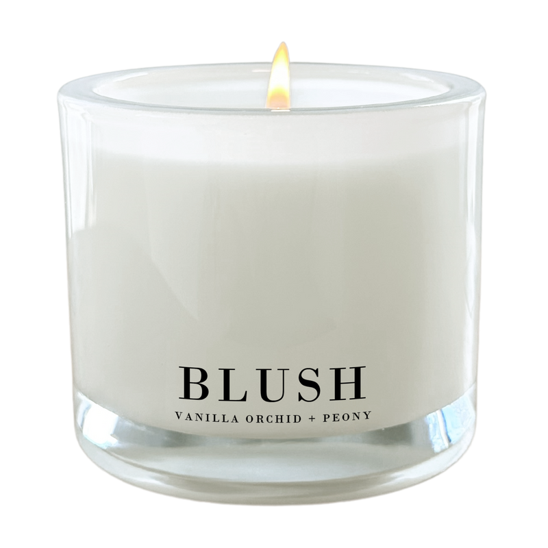 A white candle with a lit flame, labeled as "BLUSH" and containing vanilla orchid and peony scents.