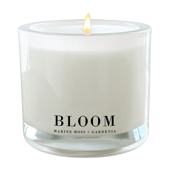 A white candle with a lit flame, labeled "Bloom" and "Marine Moss + Gardenia".