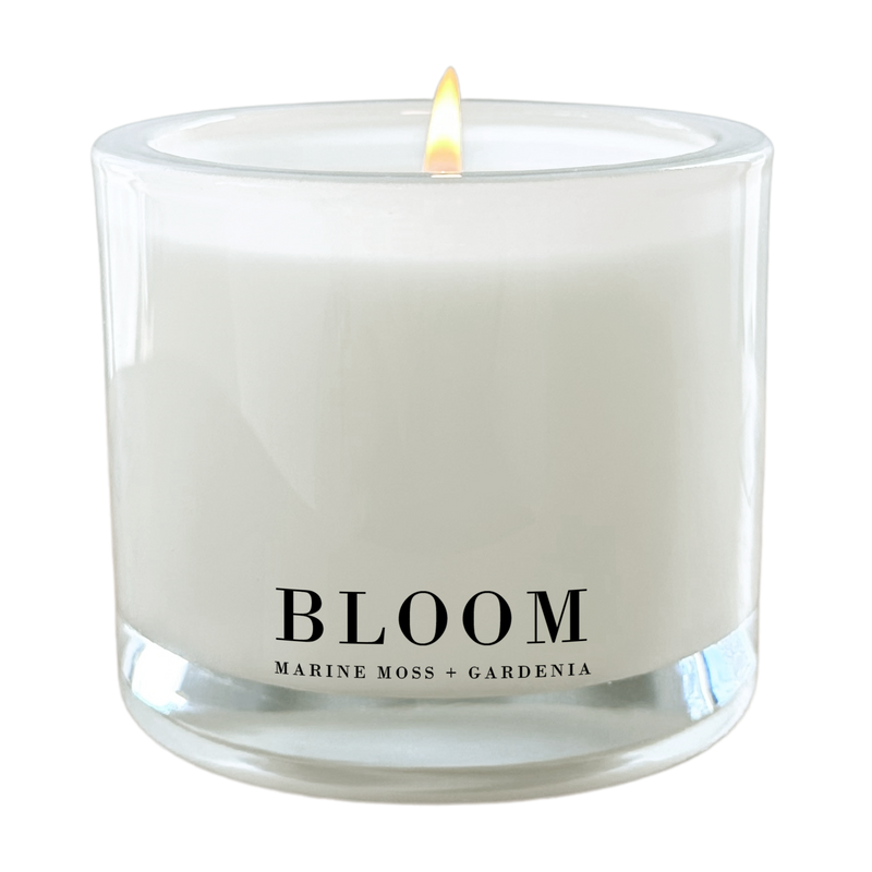 A white candle with a lit flame, labeled "Bloom" and "Marine Moss + Gardenia".