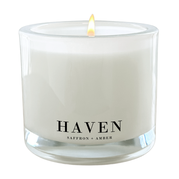 A white candle with a lit flame, labeled "HAVEN" and "SAFFRON + AMBER" on its surface.