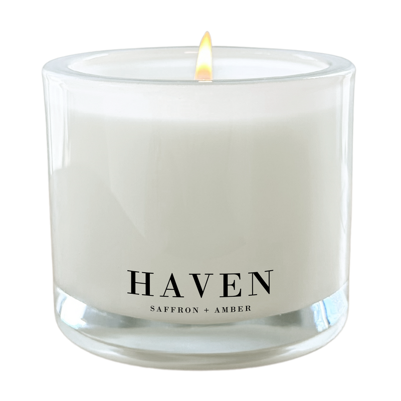 A white candle with a lit flame, labeled "HAVEN" and "SAFFRON + AMBER" on its surface.