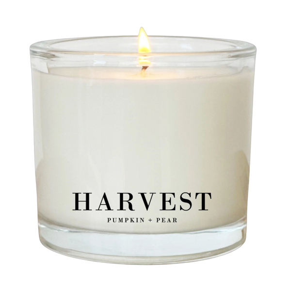 A white candle with a lit flame, labeled "Harvest Pumpkin + Pear".