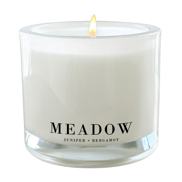 a white candle with a lit flame, labeled as a "Meadow" candle.