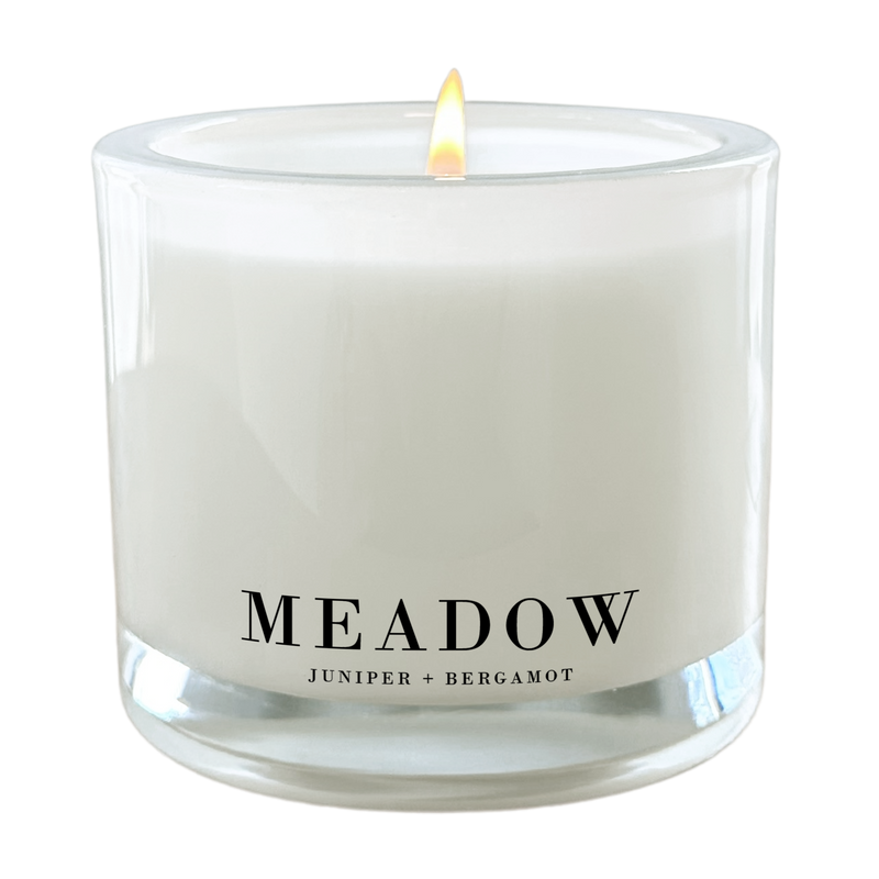 a white candle with a lit flame, labeled as a "Meadow" candle.
