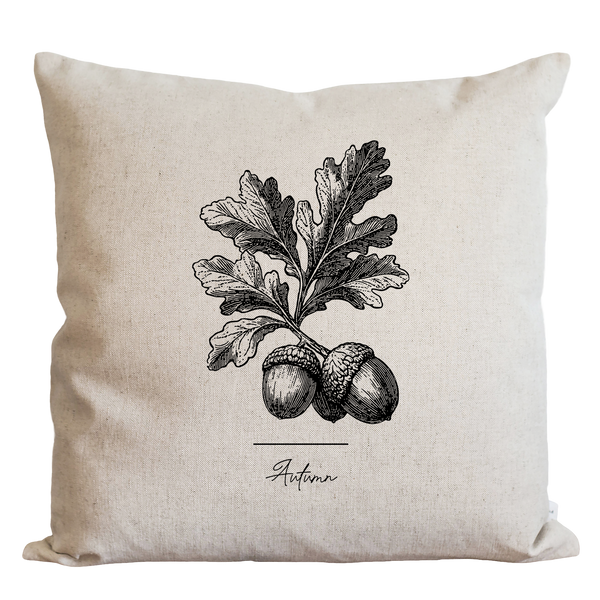 A beige pillow with a black and white illustration of an oak tree and acorns on it, along with the word "Autumn" written below the illustration.