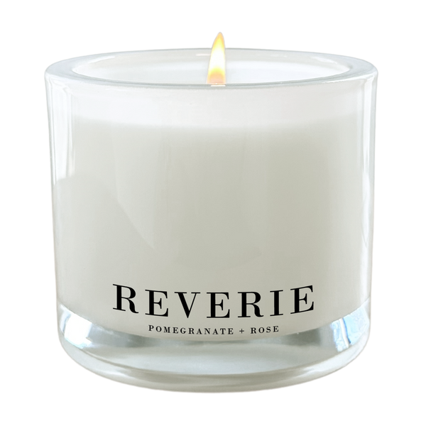 a white candle with a lit flame, labeled as "Reverie" and containing "Pomegranate + Rose" as a scent.