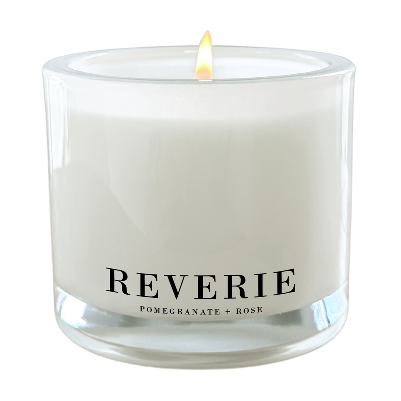 a white candle with a lit flame, labeled as "Reverie" and containing "Pomegranate + Rose" as a scent.