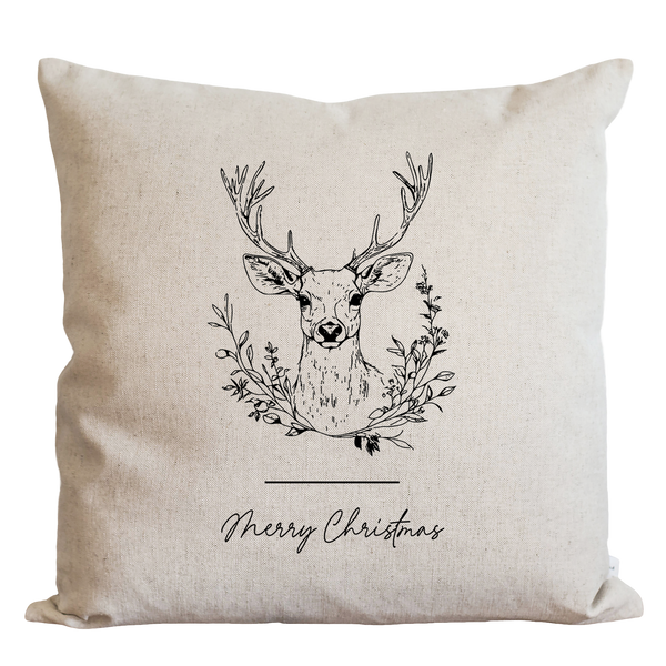 A beige pillow with a black and white illustration of a deer head surrounded by a wreath of leaves and branches, along with the text "Merry Christmas" below the image.