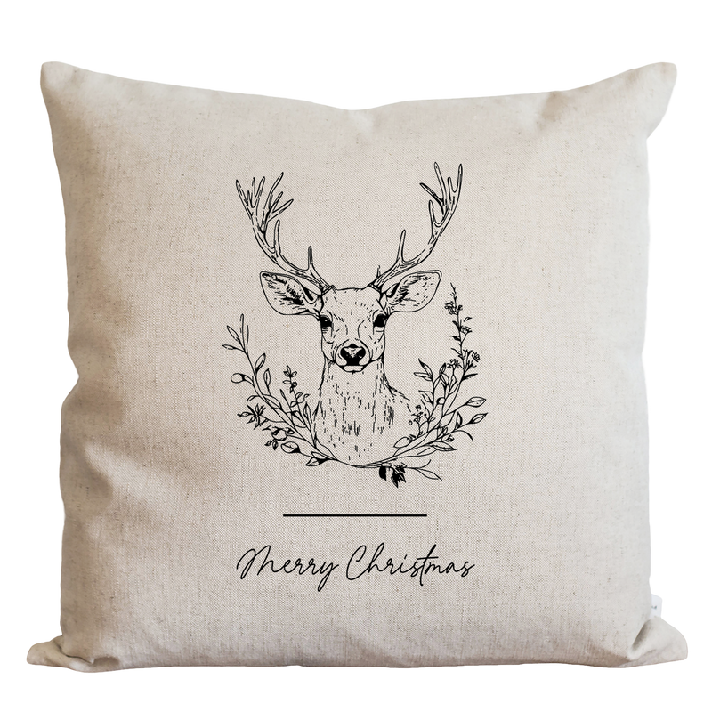 A beige pillow with a black and white illustration of a deer head surrounded by a wreath of leaves and branches, along with the text "Merry Christmas" below the image.