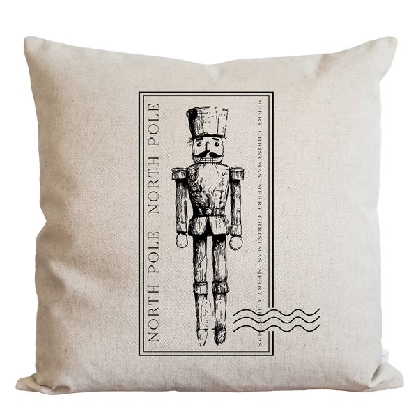 A beige pillow with a black and white illustration of a nutcracker soldier in the center, surrounded by text that reads "NORTH P Pole" and "MERRY CHRISTMAS".