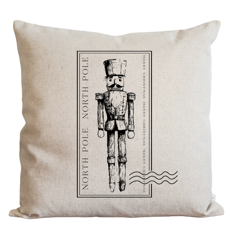 A beige pillow with a black and white illustration of a nutcracker soldier in the center, surrounded by text that reads "NORTH P Pole" and "MERRY CHRISTMAS".